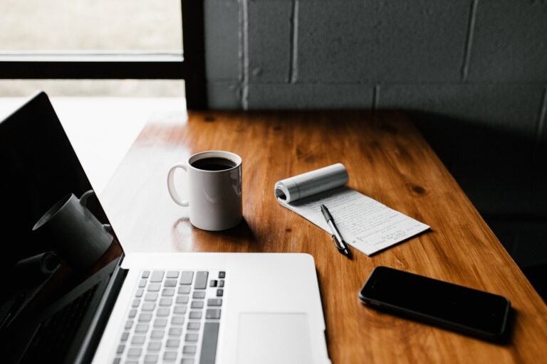 A laptop, notebook and pen beside a cup of coffee on a wooden desk