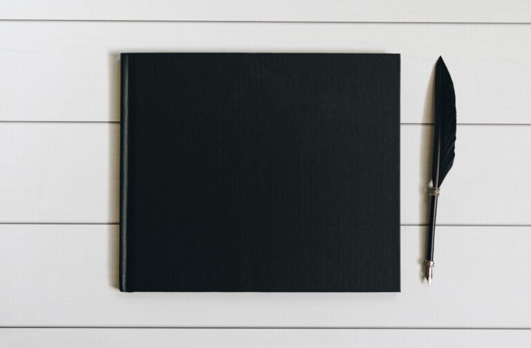 A blank black hardcover book beside a black feather quill pen