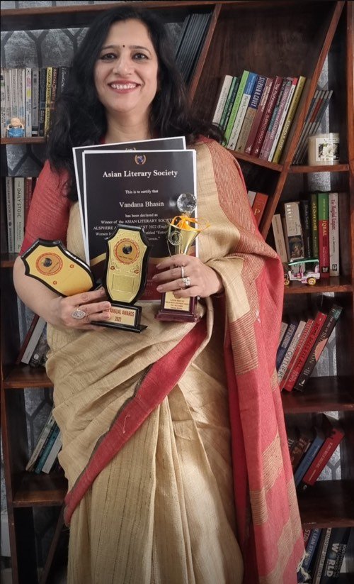 Vandana Bhasin standing at her bookshelf, holding an Asian Literary Society certificate and literary award trophies